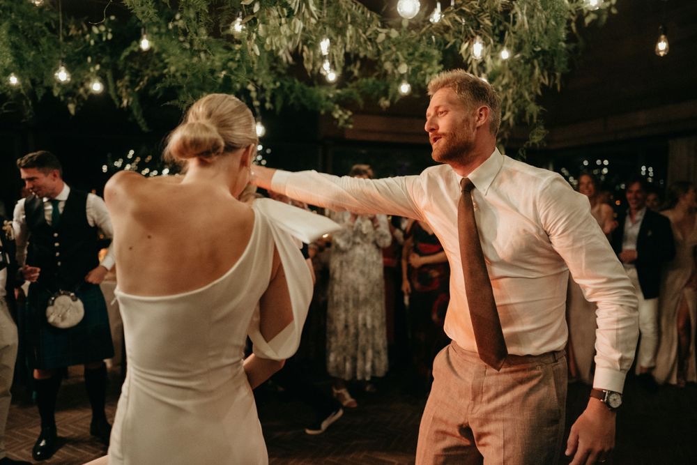 Couple dancing energetically at wedding reception under hanging garden lights.