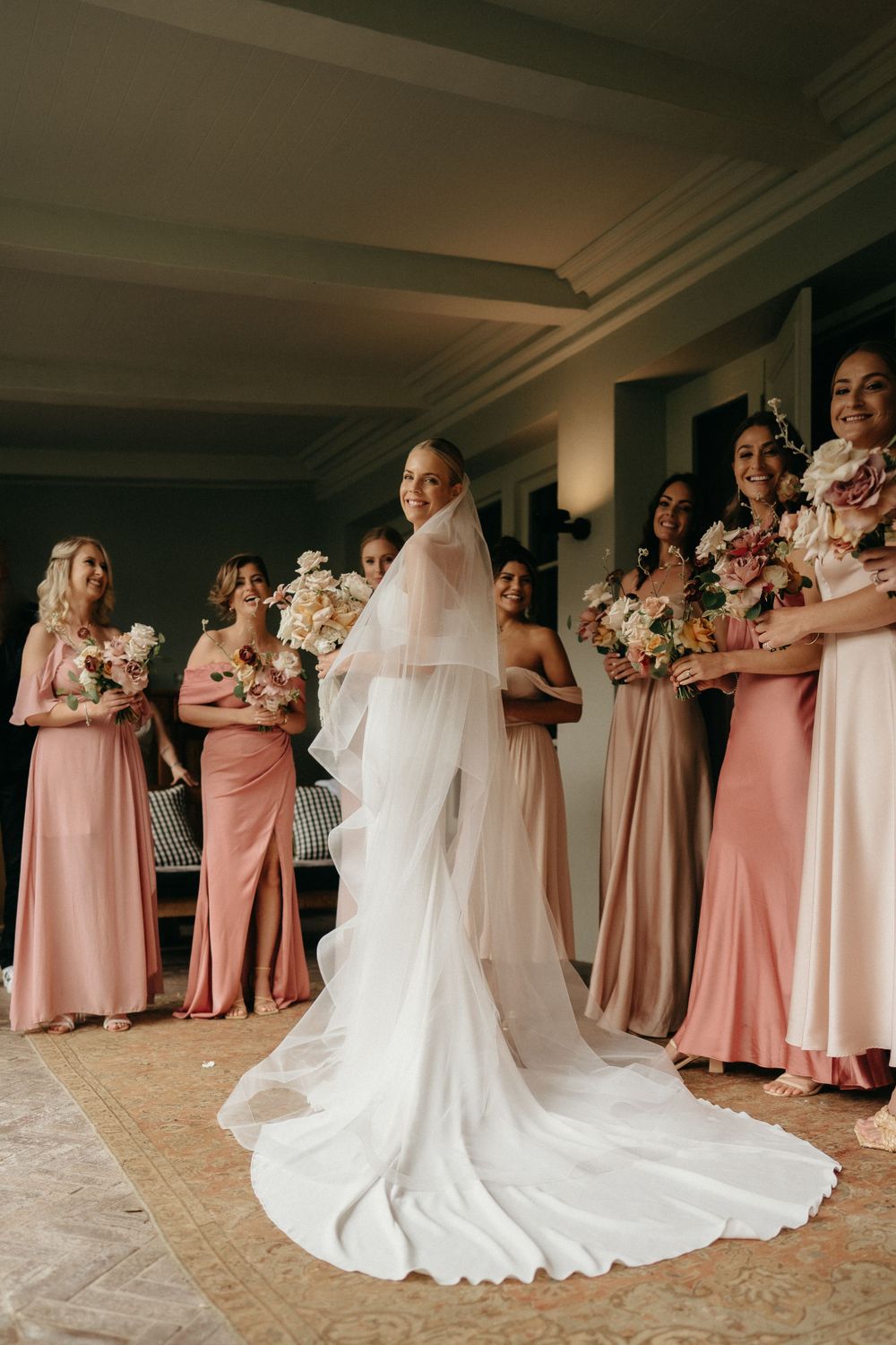 A bride in a flowing white gown stands with bridesmaids wearing blush pink dresses during a first look reveal.