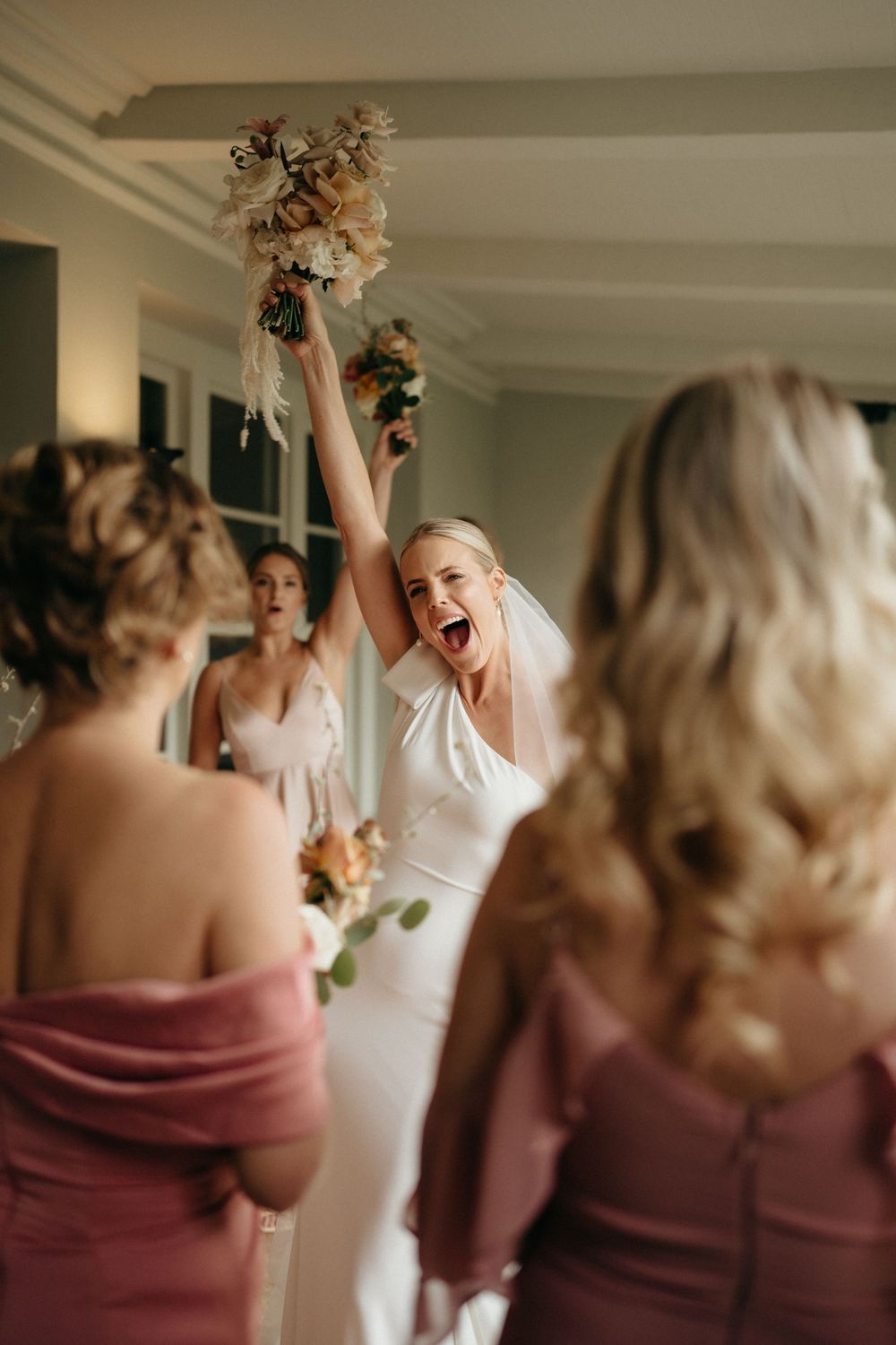 Joyful celebration moment as bride raises bouquet surrounded by bridesmaids in dusty rose dresses.