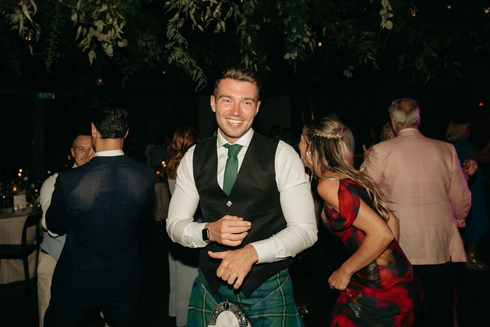 Wedding guest in traditional Scottish kilt enjoys outdoor evening reception.