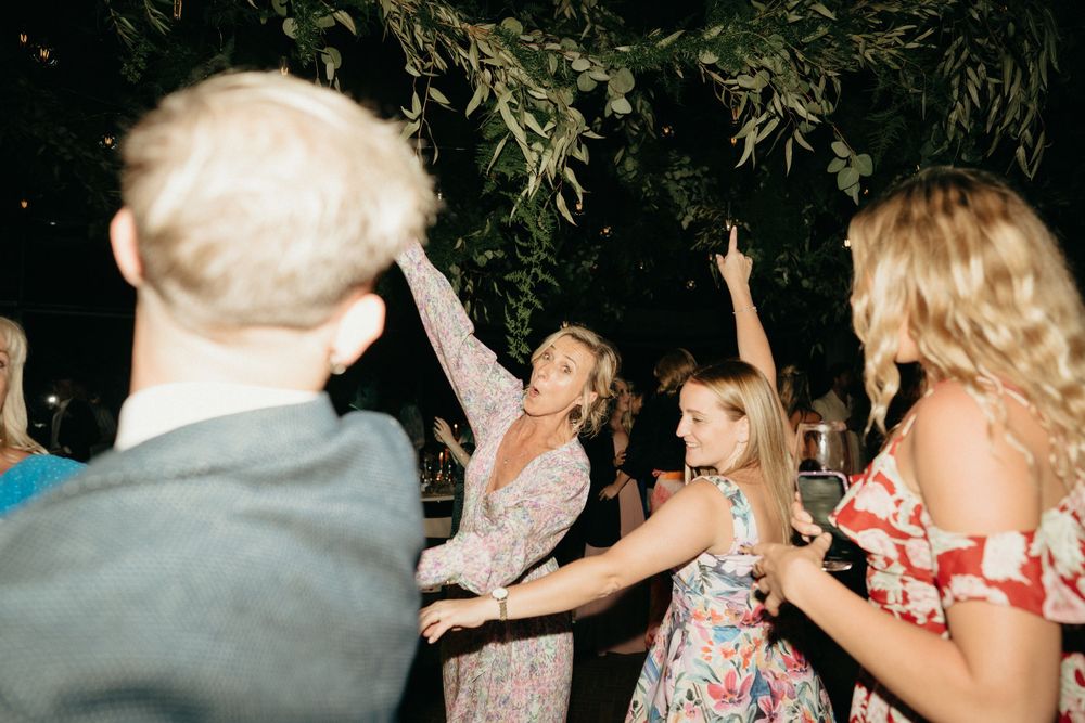 Wedding guests dancing and celebrating beneath foliage and string lights at outdoor reception.