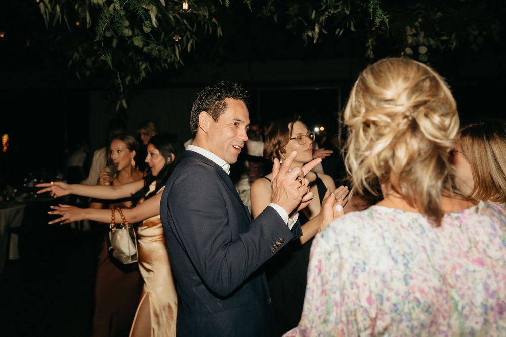 Wedding reception dance party with guests in formal attire under greenery and lights.