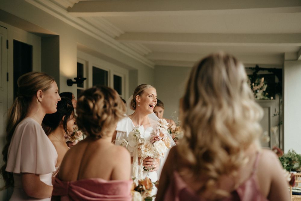 A group of women in soft-colored dresses gather together in a sunlit room, creating a warm and intimate moment.