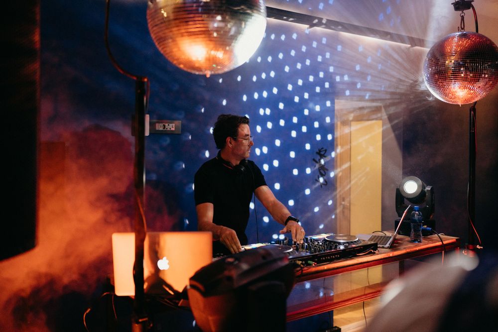 DJ performs behind decks with disco balls and atmospheric lighting creating a club-like atmosphere.