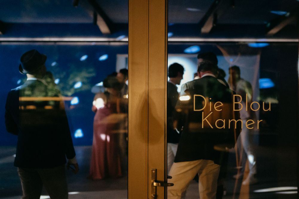 Silhouettes visible through glass door with 'Die Blou Kamer' text illuminated.