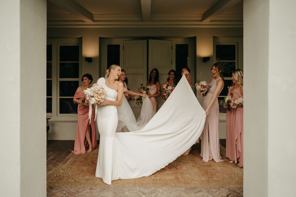 Wedding party helps adjust a flowing white bridal gown train in an elegant indoor setting.