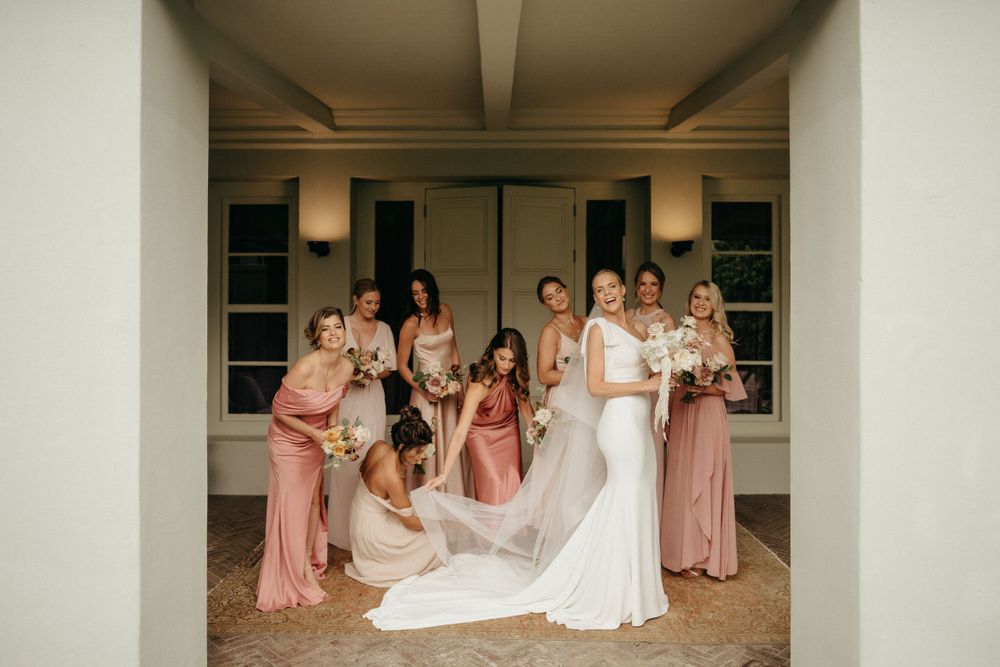 Wedding party wearing pink bridesmaid dresses poses together with bride in white gown under covered walkway.