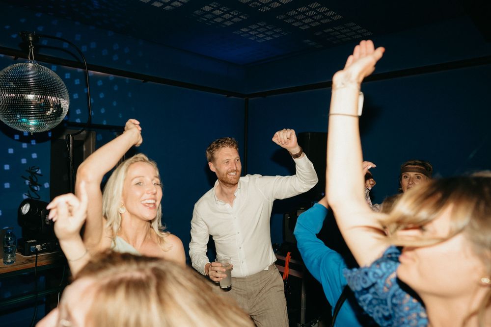Wedding guests dancing enthusiastically under a disco ball in a dark blue-lit room.