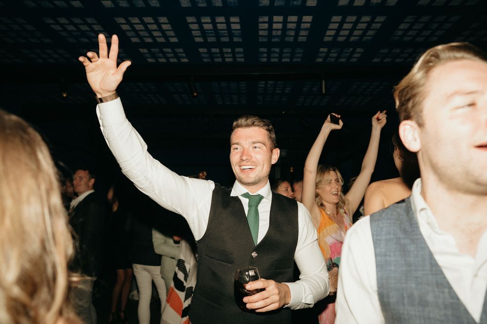 Wedding reception guests dancing and celebrating with raised arms under mood lighting.