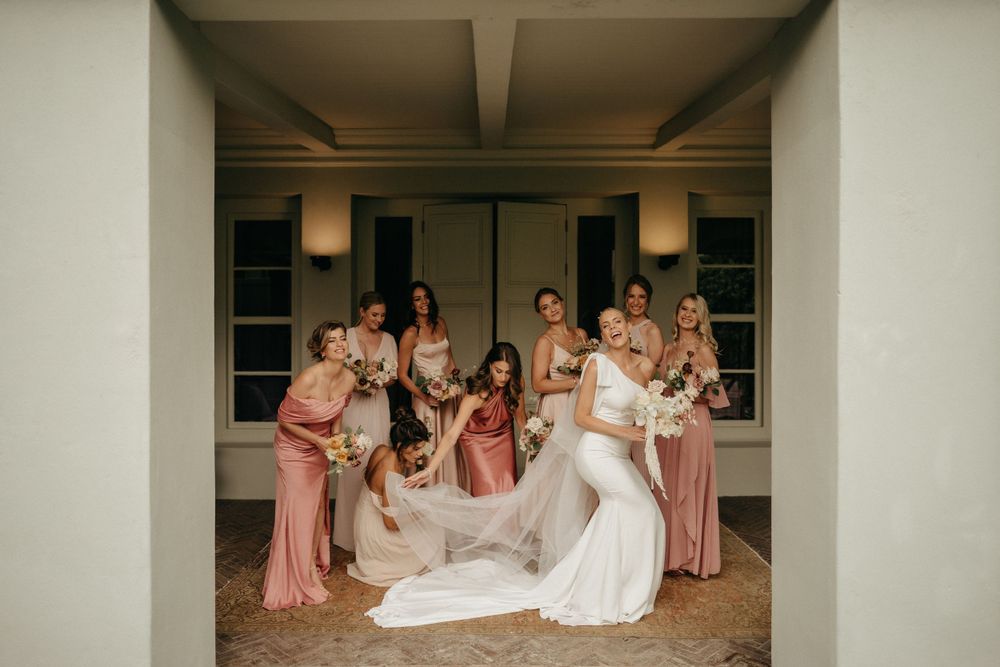 A bridal party in pink dresses gathered in an interior hallway for a celebratory photo.