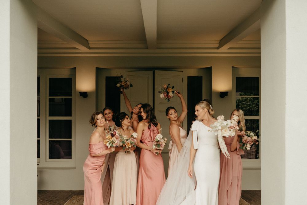 Wedding party in blush pink and white dresses celebrating with raised bouquets under covered porch.