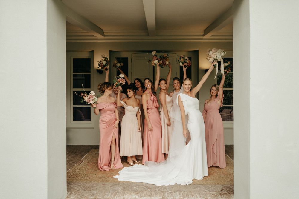 A group in pink bridesmaid dresses and a bride in white celebrate joyfully with raised bouquets in a covered walkway.