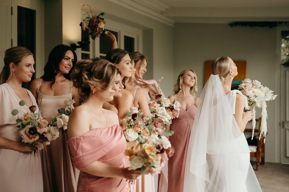 Bridesmaids in pink dresses holding floral bouquets gather around with the bride in white.
