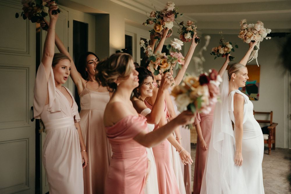 Group of bridesmaids in pink dresses lift bouquets joyfully during wedding celebration indoors.