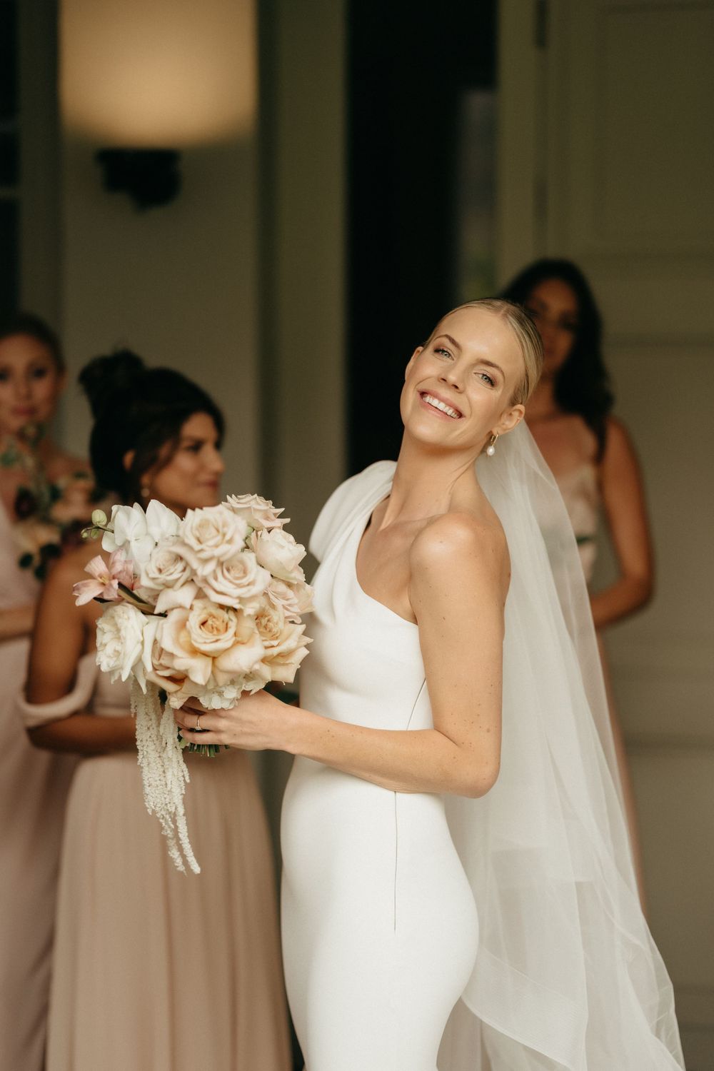A bride in a sleek white dress holds a bouquet of cream roses while wearing a flowing veil.