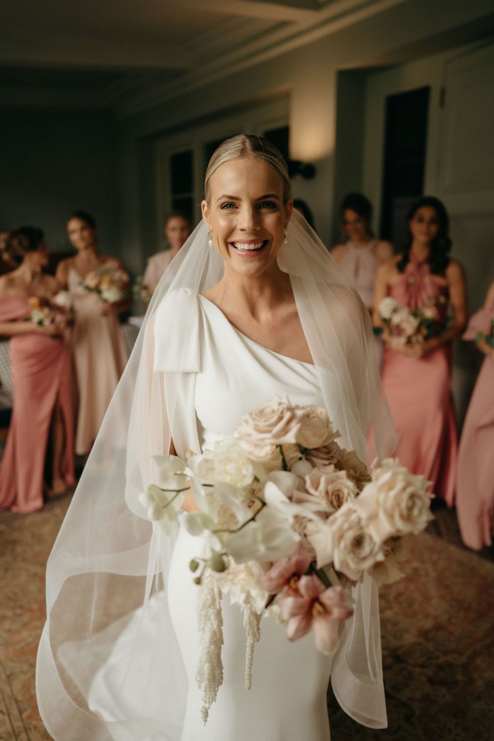 A bride in a white dress and veil holds a blush and cream rose bouquet while bridesmaids in pink dresses stand behind her.