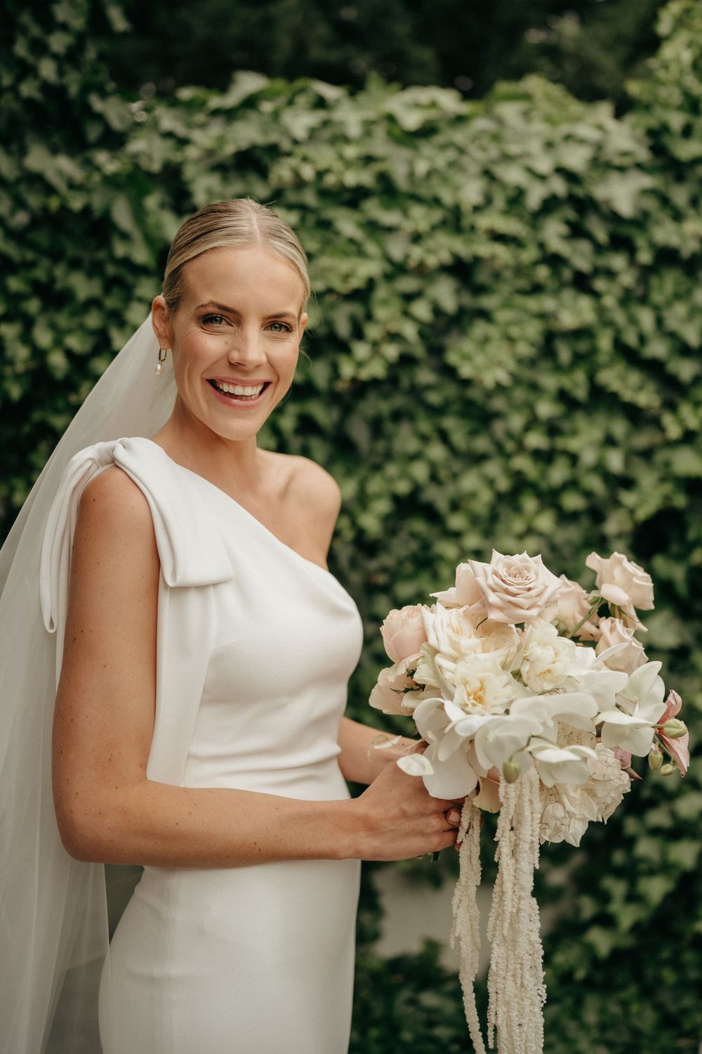 A bride in an elegant white gown holds a romantic bouquet of blush and cream flowers against an ivy backdrop.