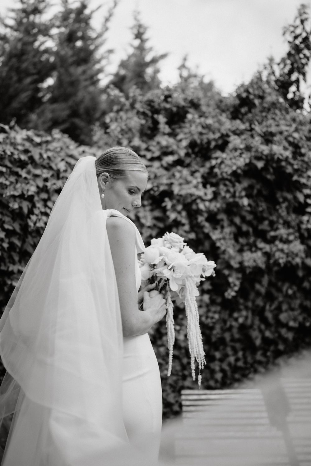 Bride in elegant white gown and veil holds bouquet of white flowers against garden backdrop.