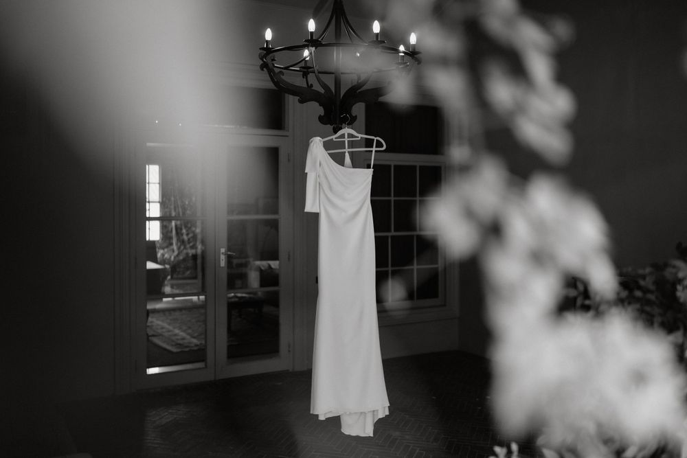 A white wedding dress hanging from chandelier in moody black and white lighting.