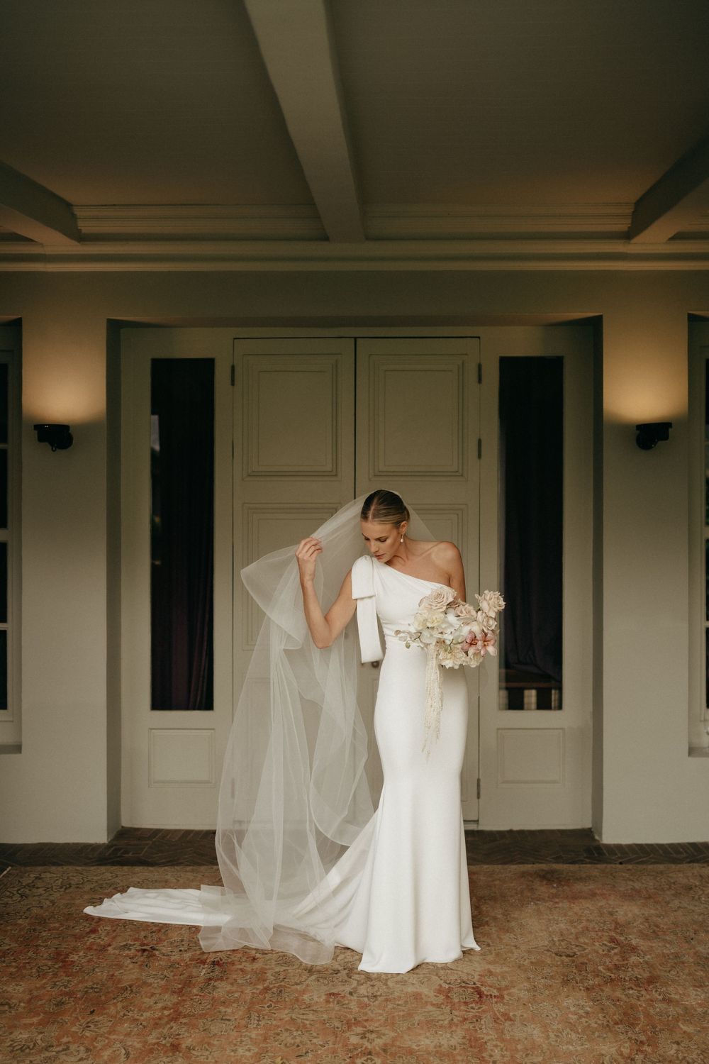 A bride in an elegant white wedding gown and long flowing veil stands gracefully against double doors with wall sconces.