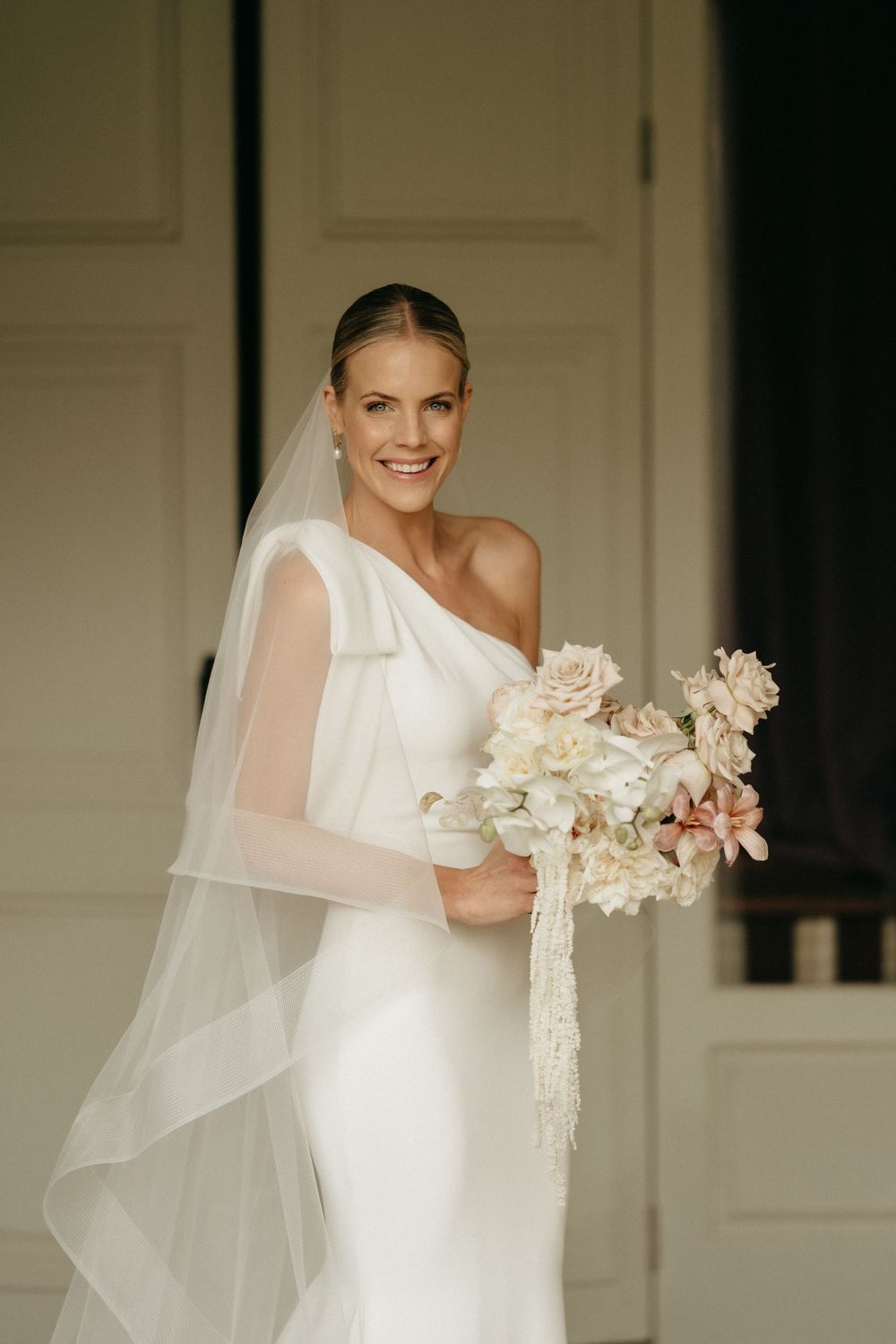 Elegant bride in one-shoulder white gown and veil holding romantic blush and cream floral bouquet.