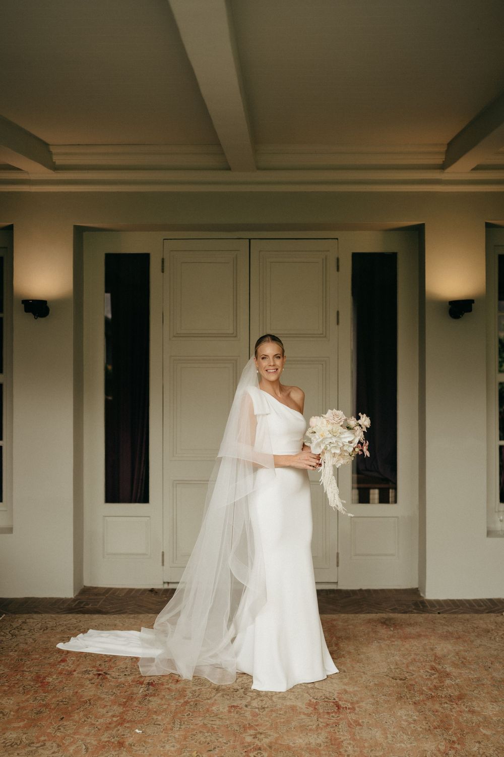 Classic bridal portrait with white one-shoulder gown and veil against neutral architectural backdrop.