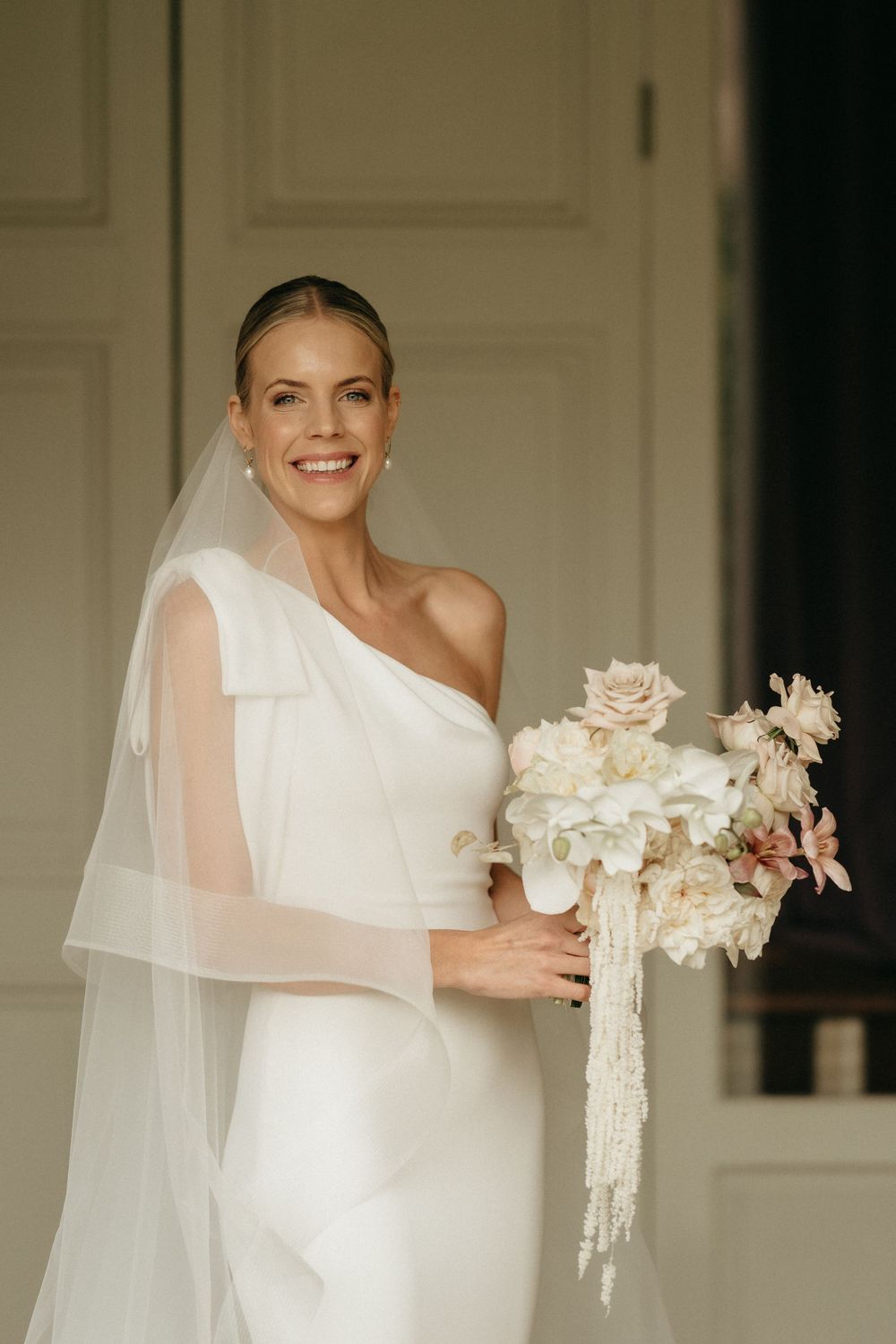 Elegant bride in one-shoulder white gown holding delicate bouquet of roses and orchids.