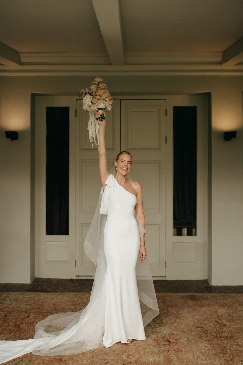 A bride in a one-shoulder white wedding gown with a long train holds her bouquet high in celebration.