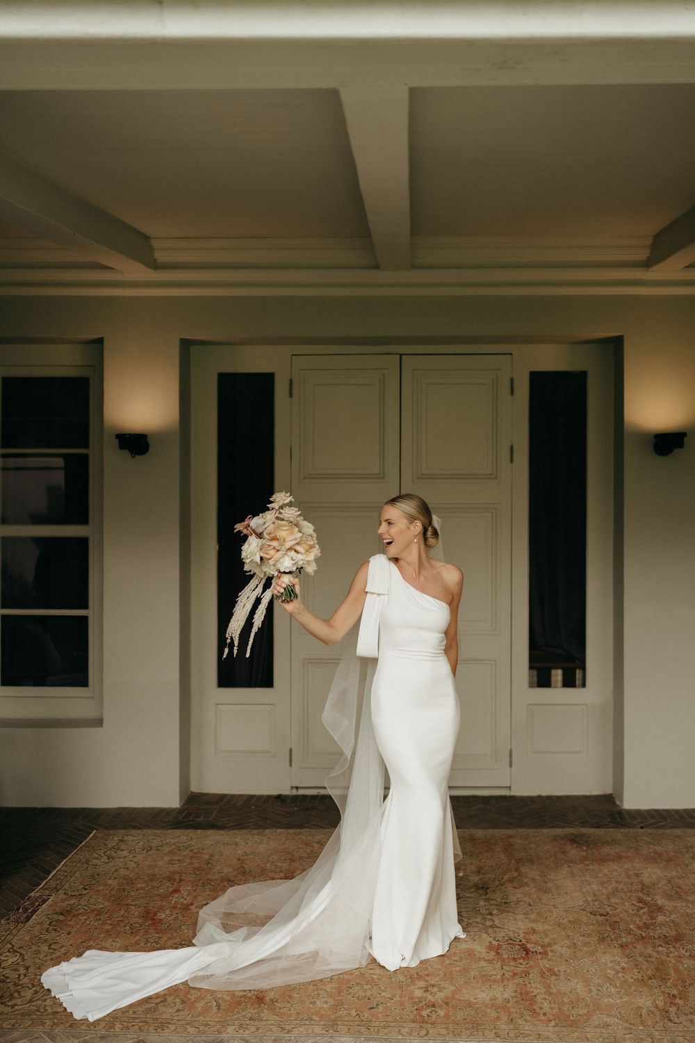A person in a one-shoulder white wedding gown with a flowing train holds a bouquet on an elegant covered porch.