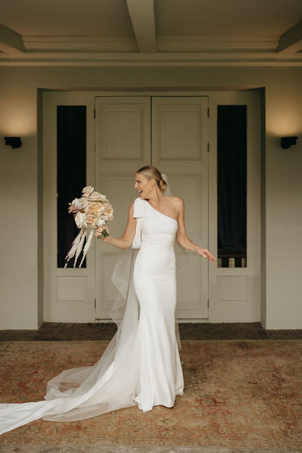 An elegant one-shoulder white wedding gown with a flowing train photographed against classic double doors.