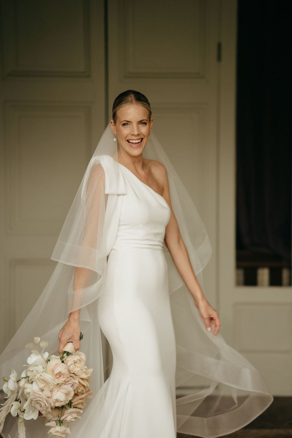 Elegant one-shoulder white wedding dress with flowing veil and neutral bouquet.