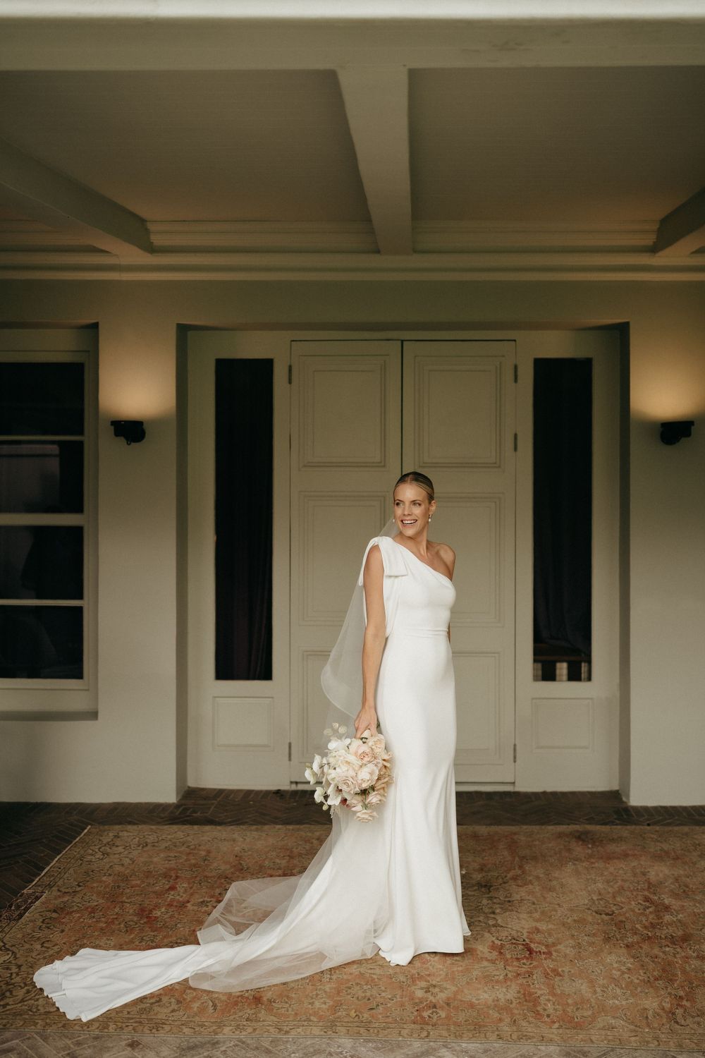 Bride poses in one-shoulder white wedding gown with train against neutral building entrance.