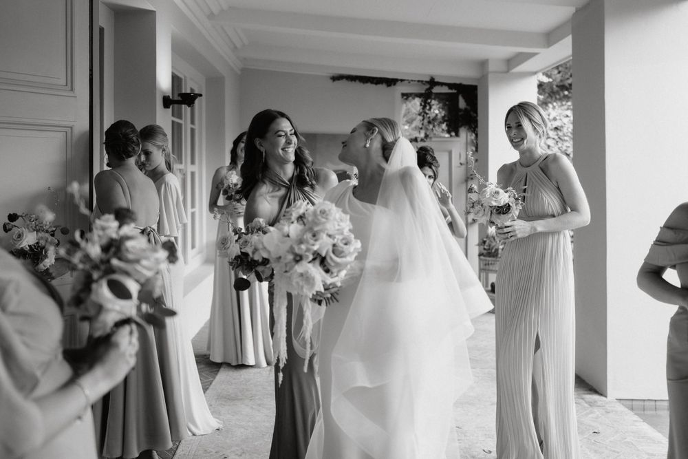 Wedding party gathered in a hallway wearing elegant gowns and holding beautiful floral bouquets captured in black and white.