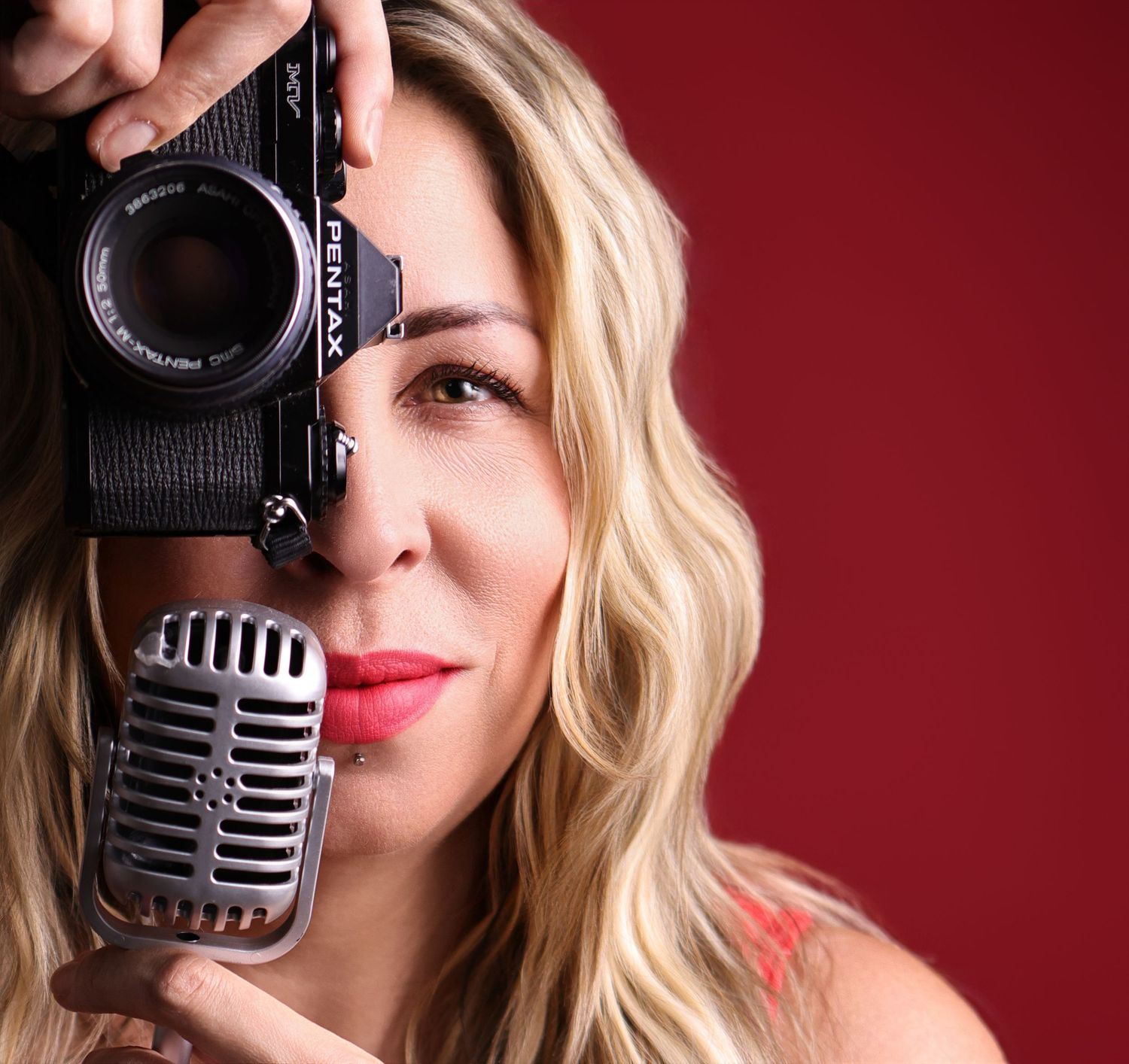 Content creator holds microphone and camera while posing against vibrant red backdrop.