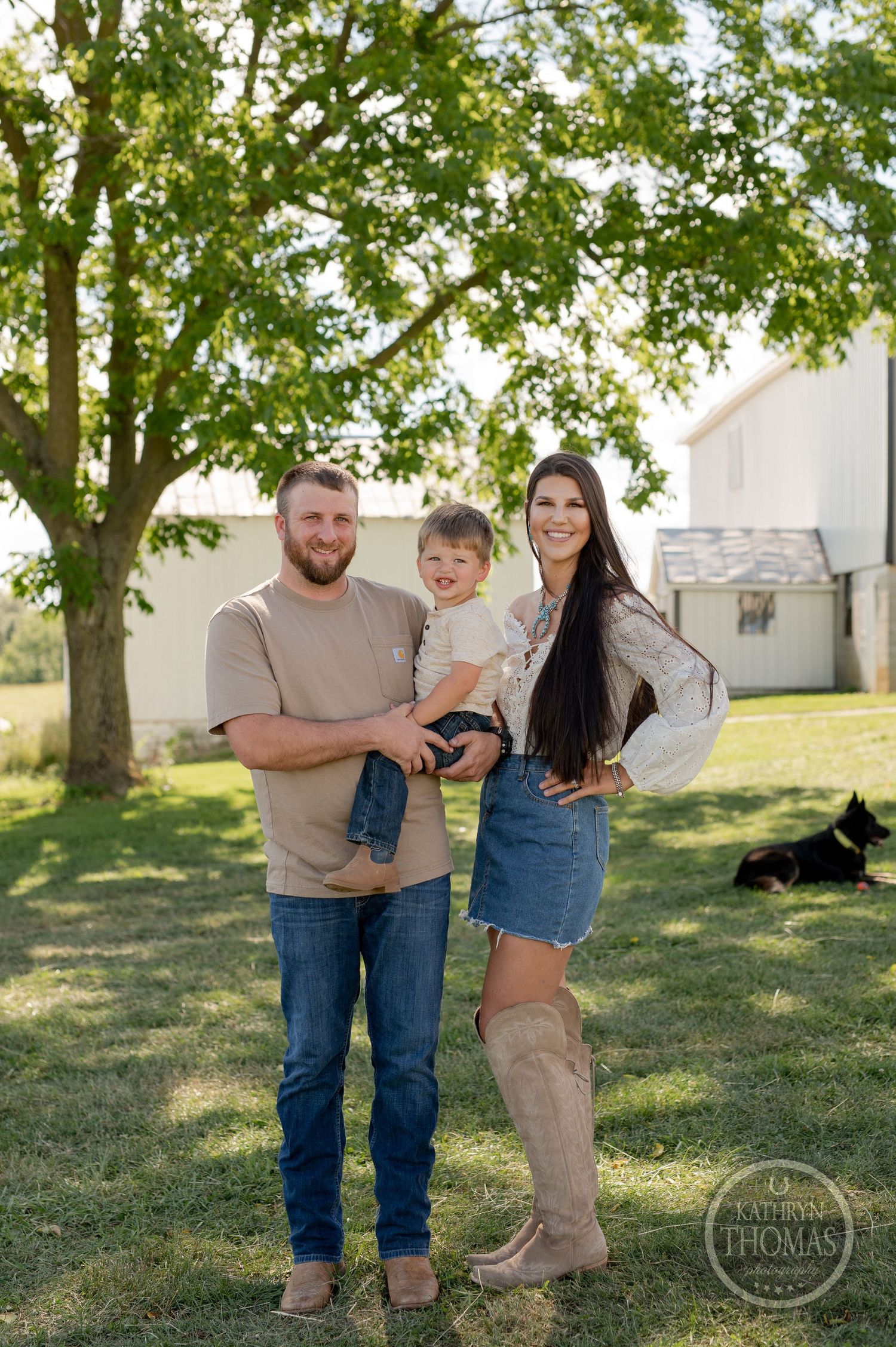 A happy family stands together outdoors with green trees and farm buildings in the background.