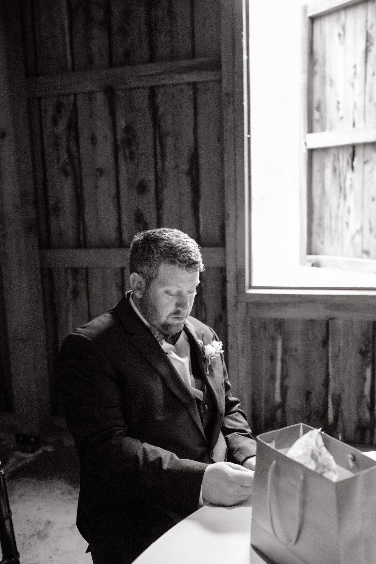 Groom in black tuxedo reads heartfelt letter before wedding ceremony in rustic barn venue.