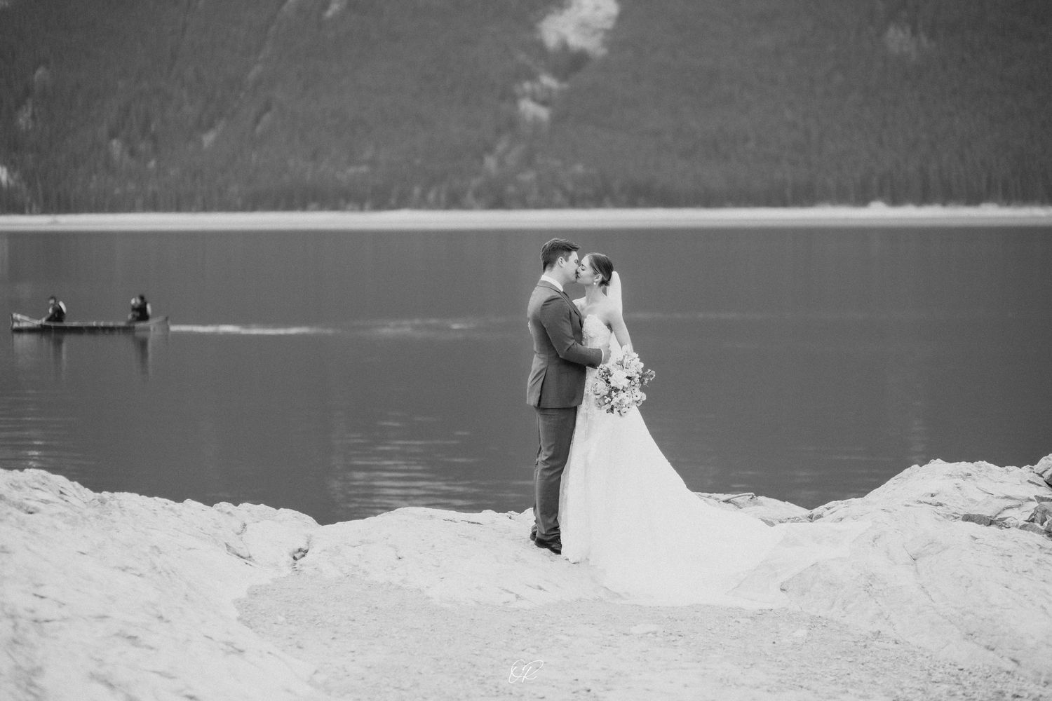 Black and white wedding portrait by a tranquil lake with mountains in background.