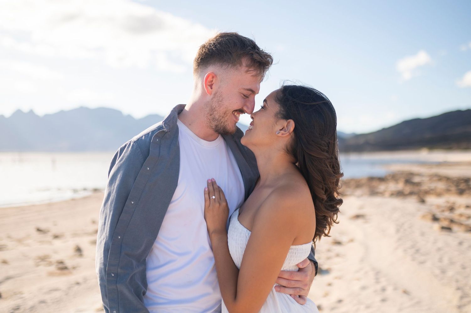 Romantic couple embracing and kissing on sandy beach with mountains in background during sunny day.