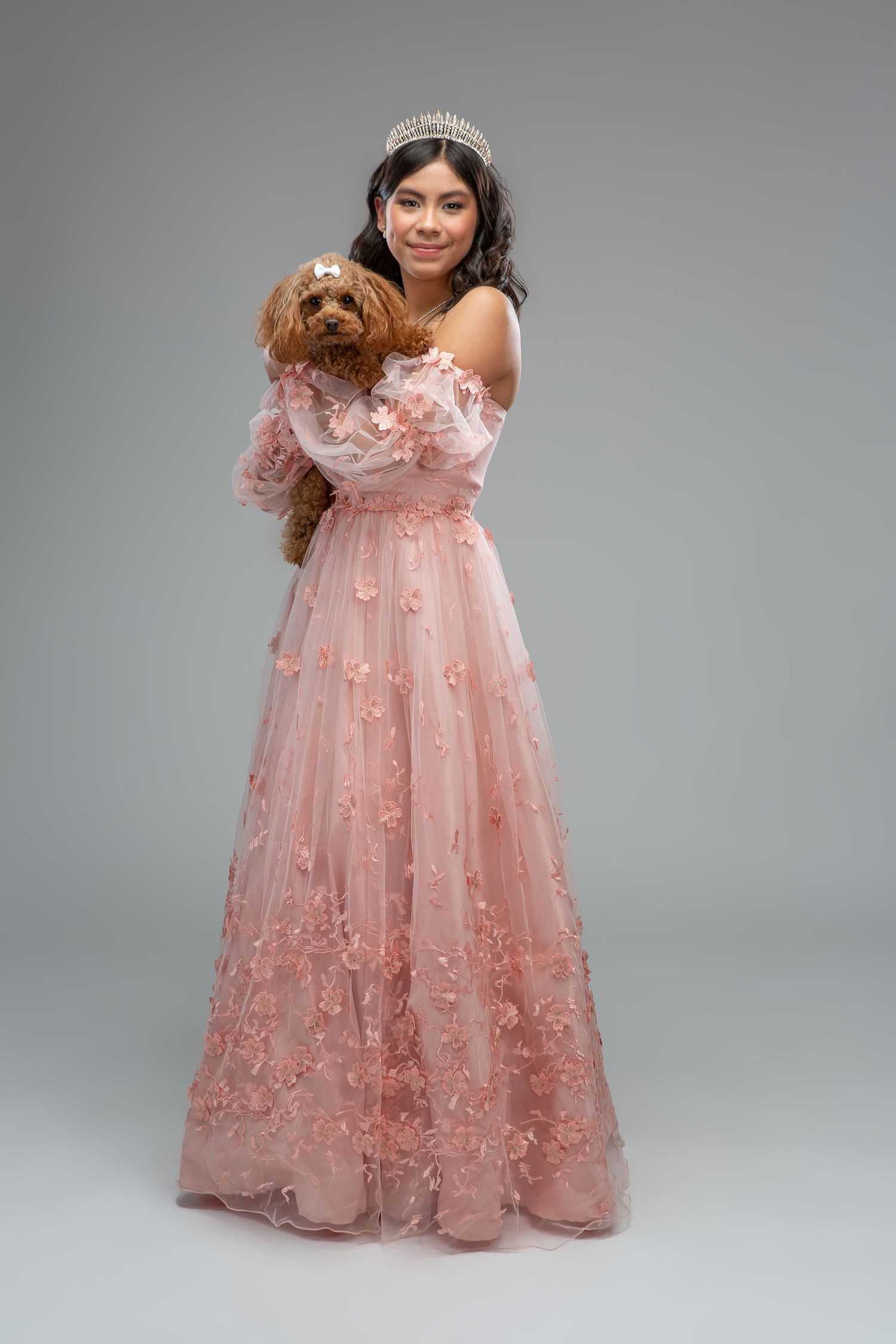 A model wears a romantic pink tulle ball gown with floral appliques and off-shoulder design against a gray backdrop.