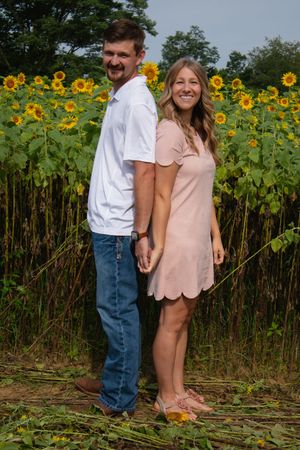 A couple poses together against a backdrop of tall blooming sunflowers on a summer day.