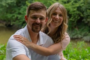 A couple embracing outdoors in a natural green setting wearing casual white clothing.