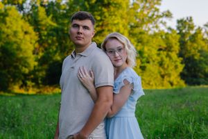 Romantic couple embraces in a sunny field with lush green foliage in the background.