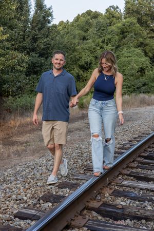 A couple walks and poses playfully along railroad tracks on a sunny day.