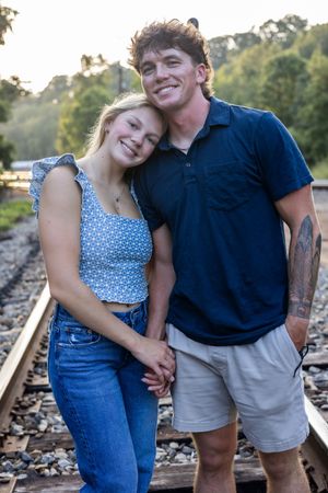 A happy couple in casual summer outfits poses together on railroad tracks during golden hour.
