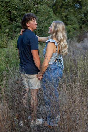 A couple holds hands while standing in tall grass near railroad tracks during sunset.
