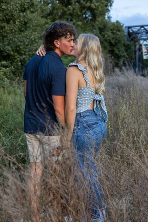 A romantic couple stands close together among tall grass with a railway bridge in the background.