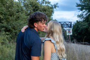 A couple shares an intimate moment near railroad tracks with a bridge visible in the background.