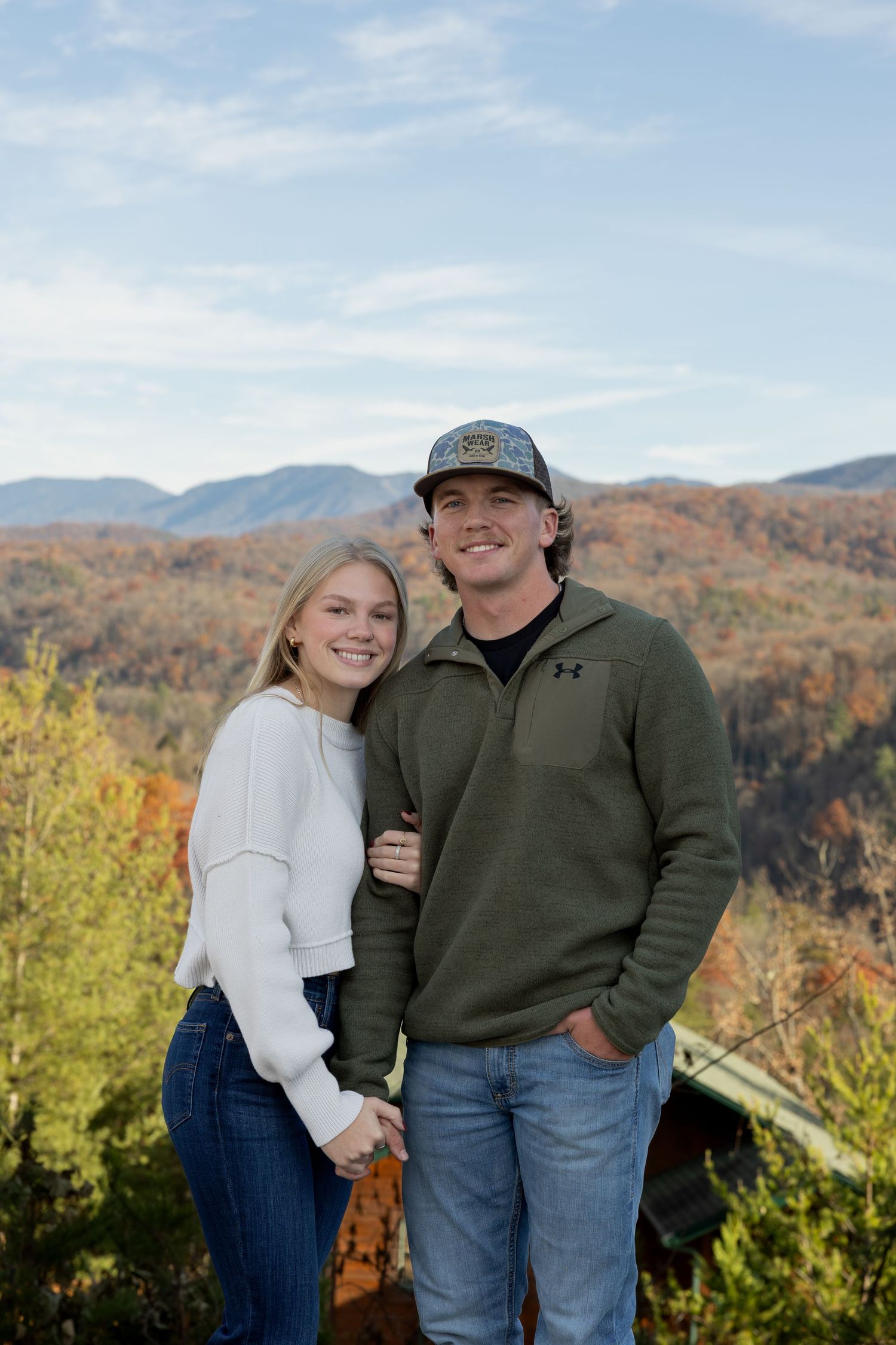 Couple embraces while standing on overlook with fall mountain landscape behind them.
