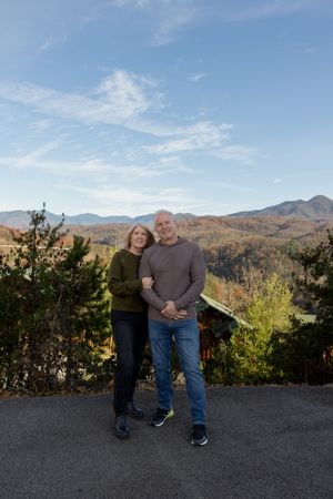 Couple poses casually on mountain viewpoint with autumn foliage in background.