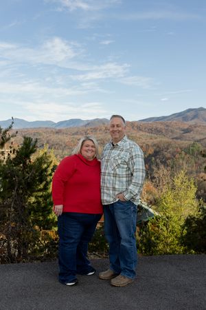 Couple stands together on overlook with autumn mountain scenery behind them.